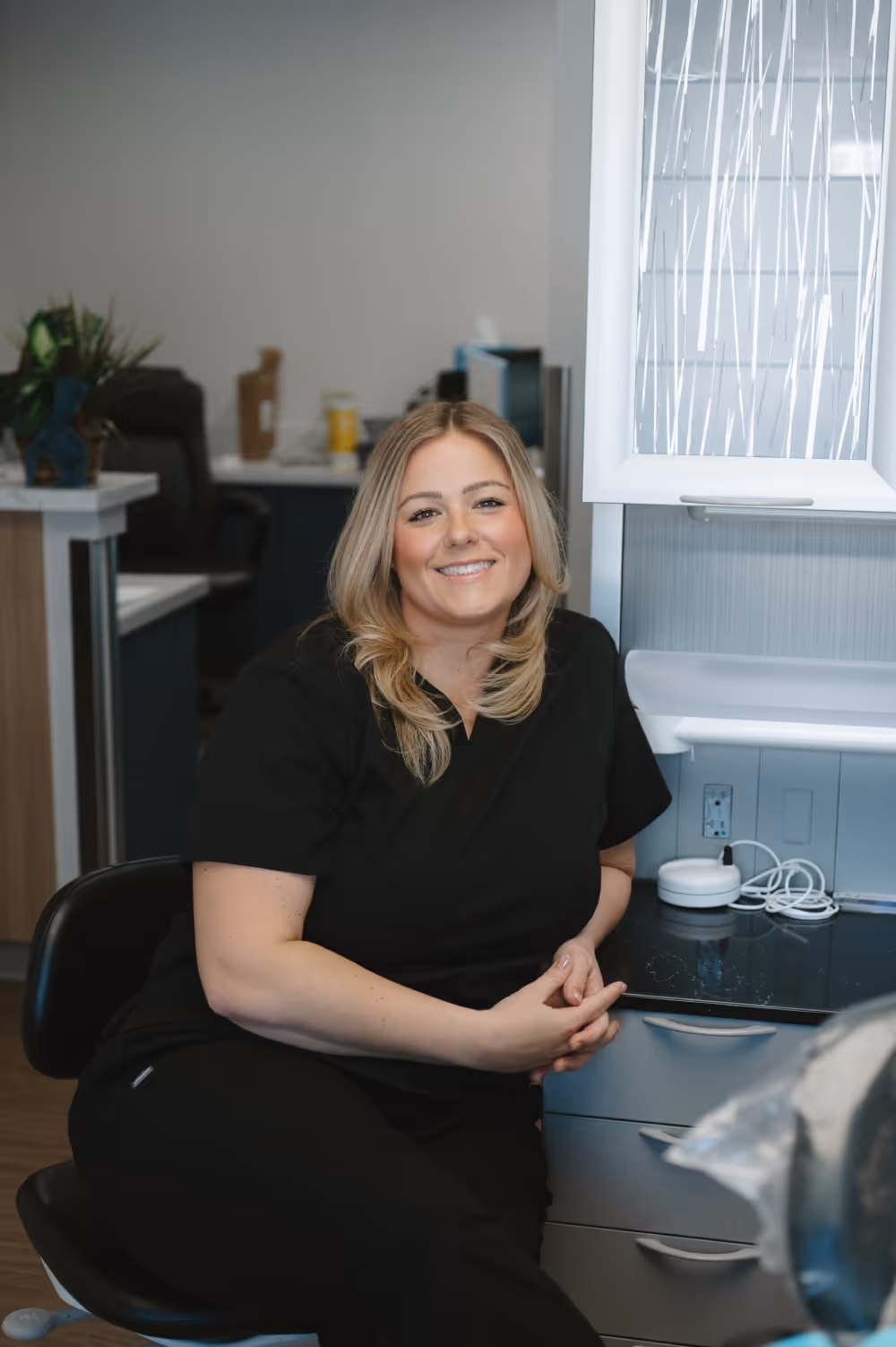 Smiling woman with blonde hair sitting on a black stool in a modern office setting, wearing black scrubs.