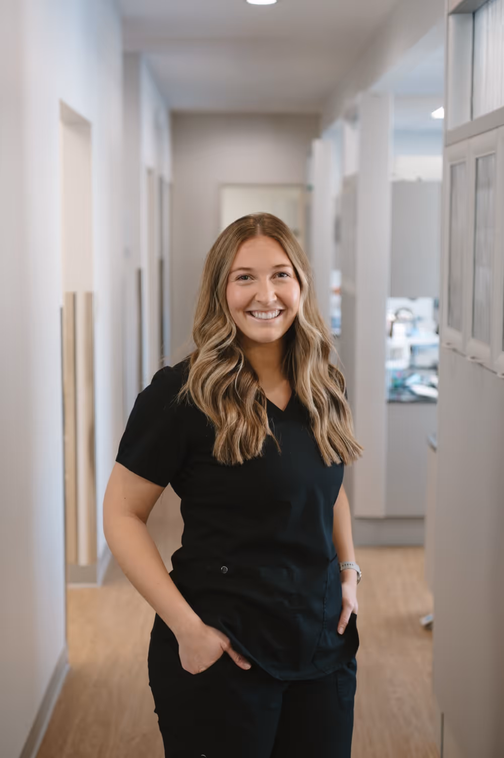 Smiling woman with long wavy hair wearing black scrubs standing in a bright hallway with hands in pockets.