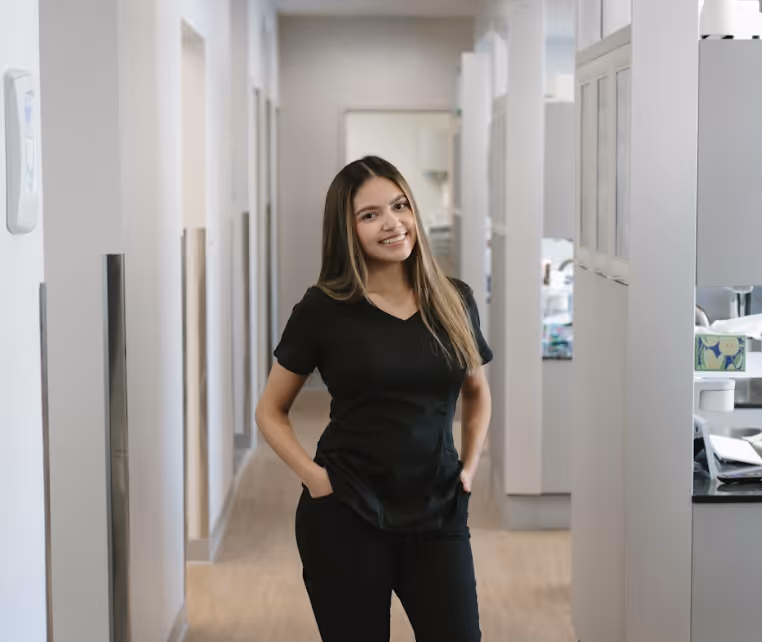 Smiling woman in black medical scrubs standing with hands in pockets in a bright hallway.