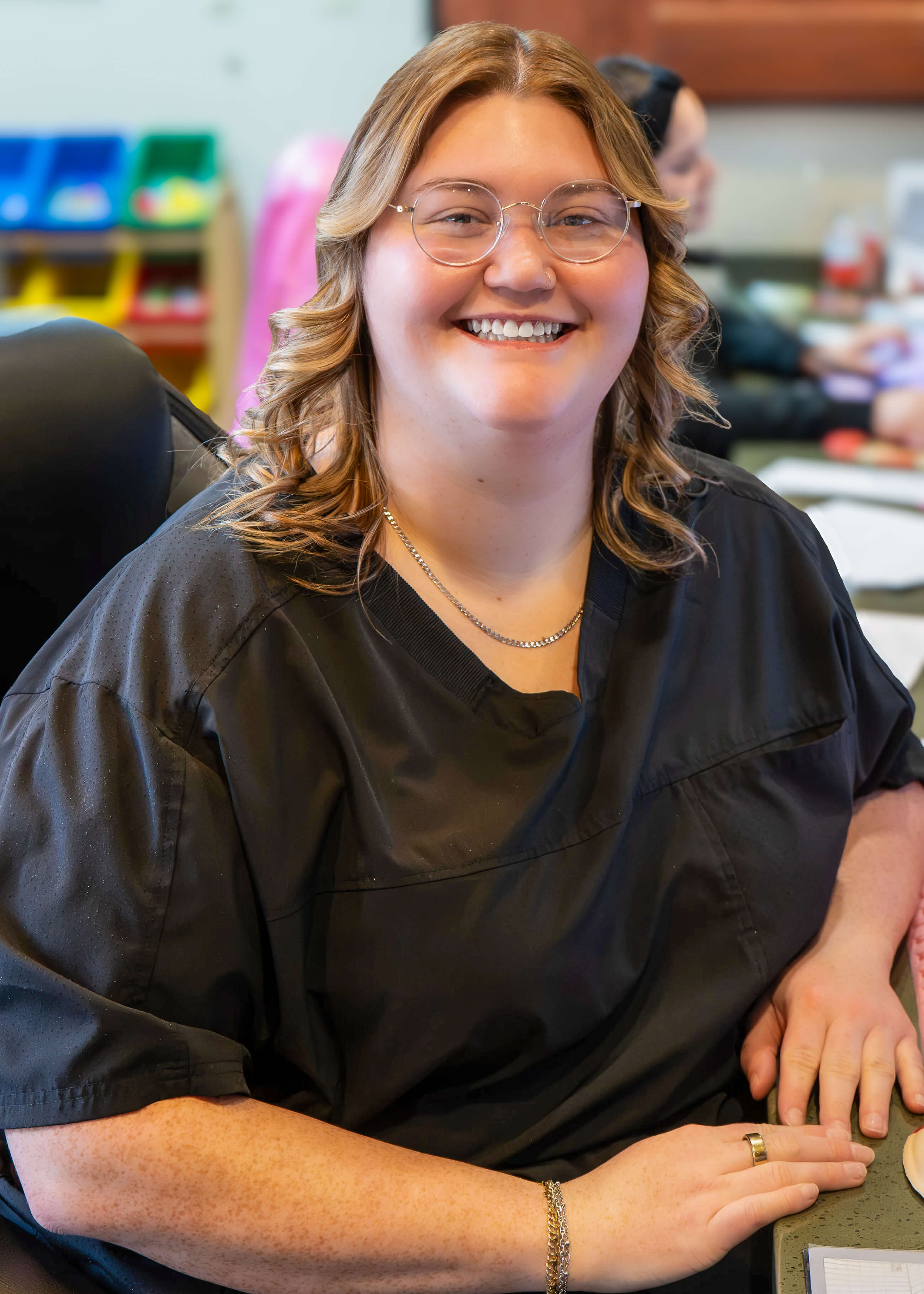 Smiling woman with glasses and curly hair wearing a black shirt sitting at a desk.