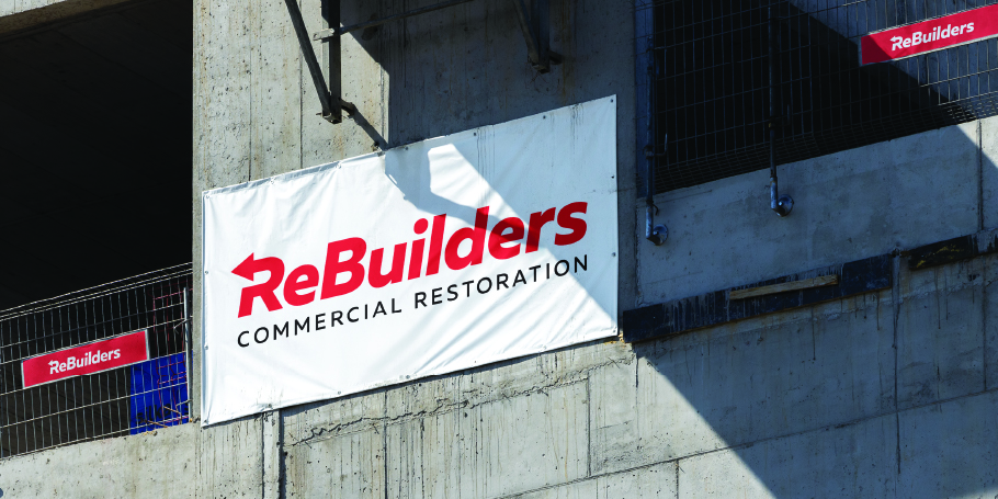 White banner with red text 'ReBuilders Commercial Restoration' hanging on a concrete wall at a construction site.