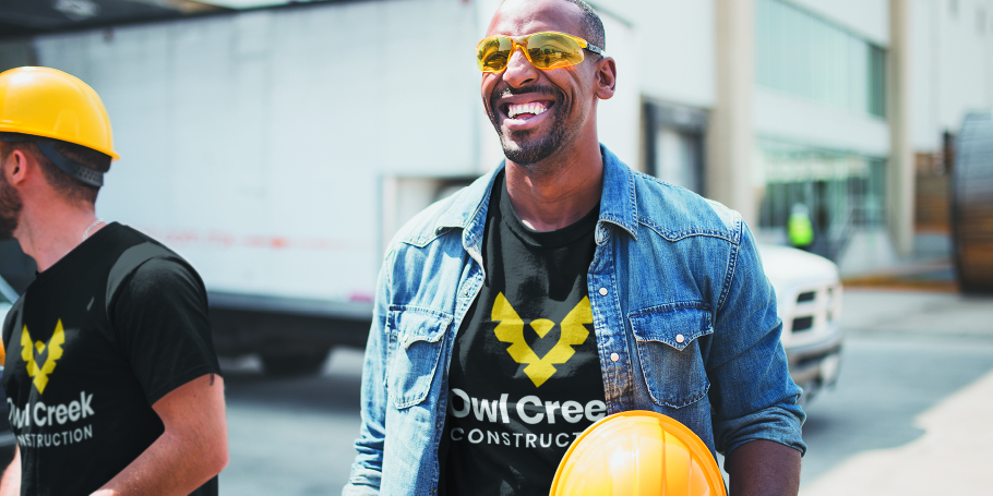 Smiling construction worker wearing yellow safety glasses and holding a yellow hard hat, dressed in an Owl Creek Construction t-shirt and denim jacket.