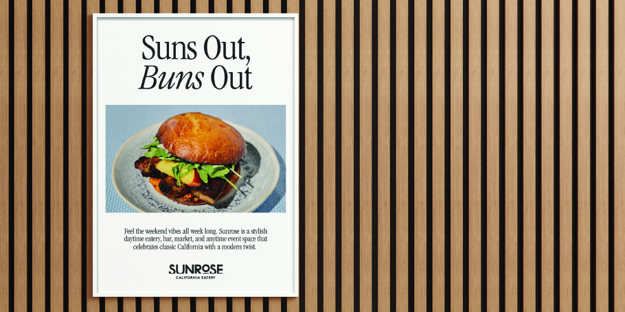 Framed poster on wooden slat wall featuring a sandwich with lettuce and tomato on a bun, titled 'Suns Out, Buns Out' promoting Sunrose California Eatery.