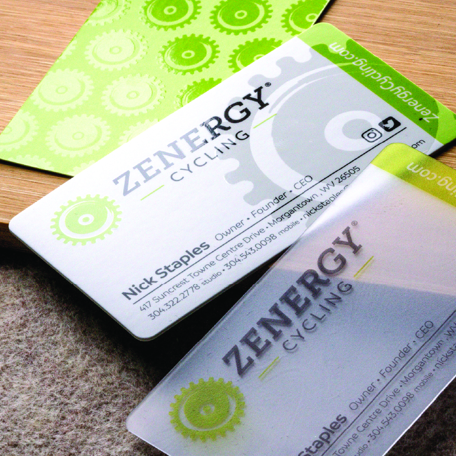 Three Zenergy Cycling business cards with green gear design and contact details for Nick Staples, Owner and CEO, on a wooden and fabric surface.