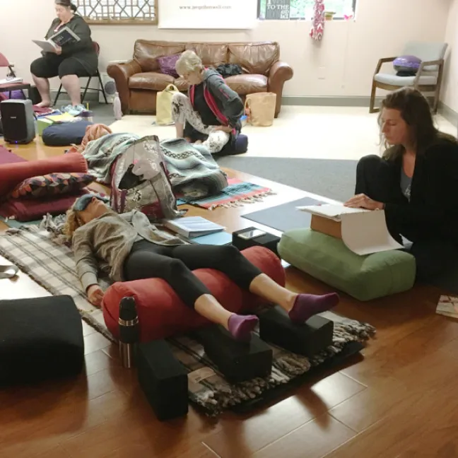 A yoga student lying on her back with blankets and another student reading to her