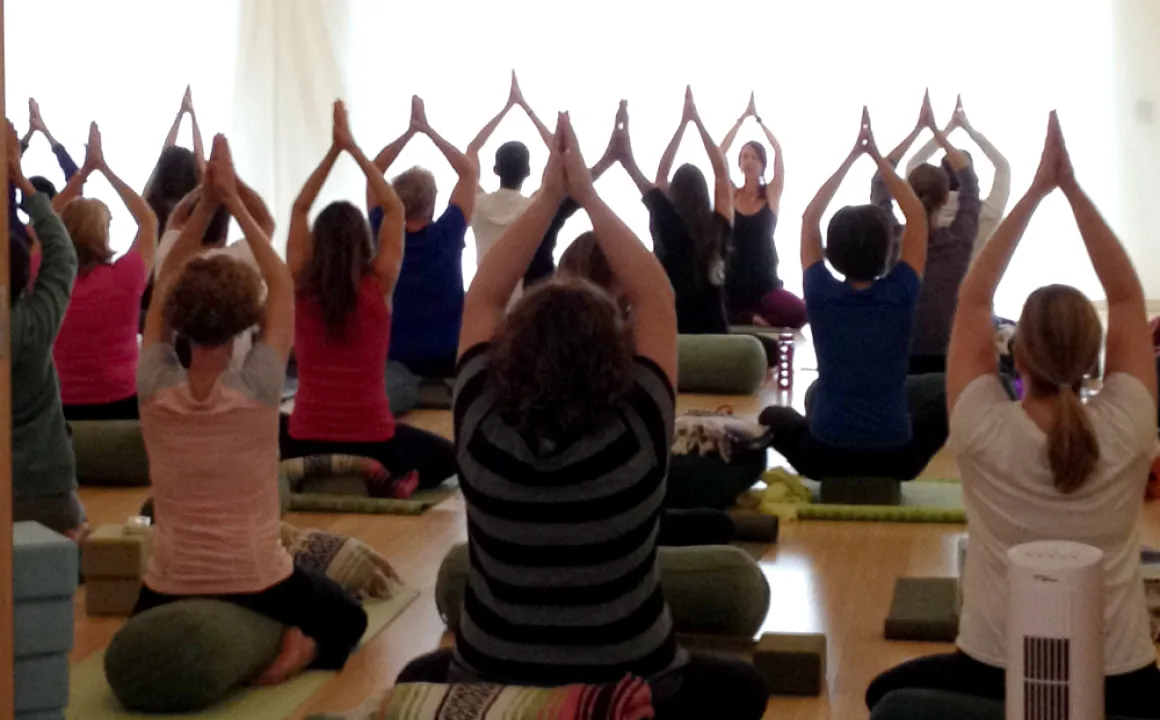 A group of people with their hands overhead in prayer position sitting on yoga mats in a yoga studio