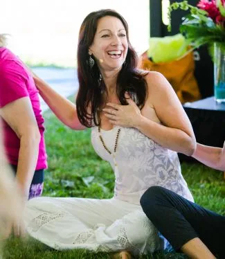 A smiling female sitting on a yoga mat with her hand on her heart in a circle with others on a mat on the grass