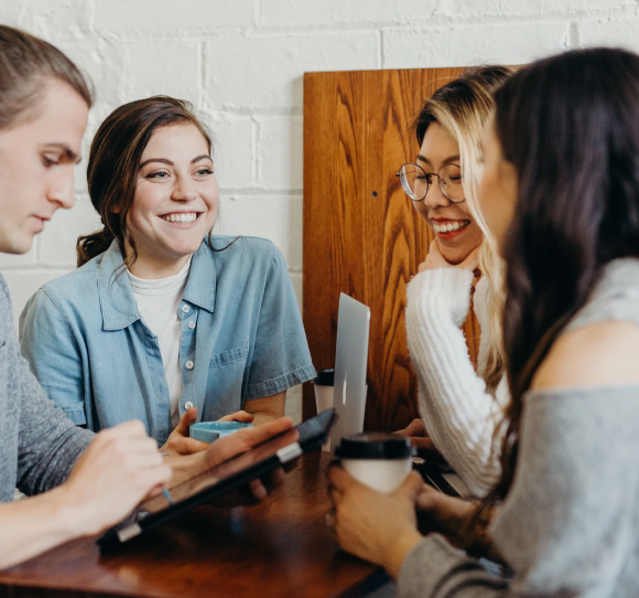 A group of four young adults sitting around a wooden table, engaging in conversation and smiling, with one person using a tablet and others holding coffee cups.