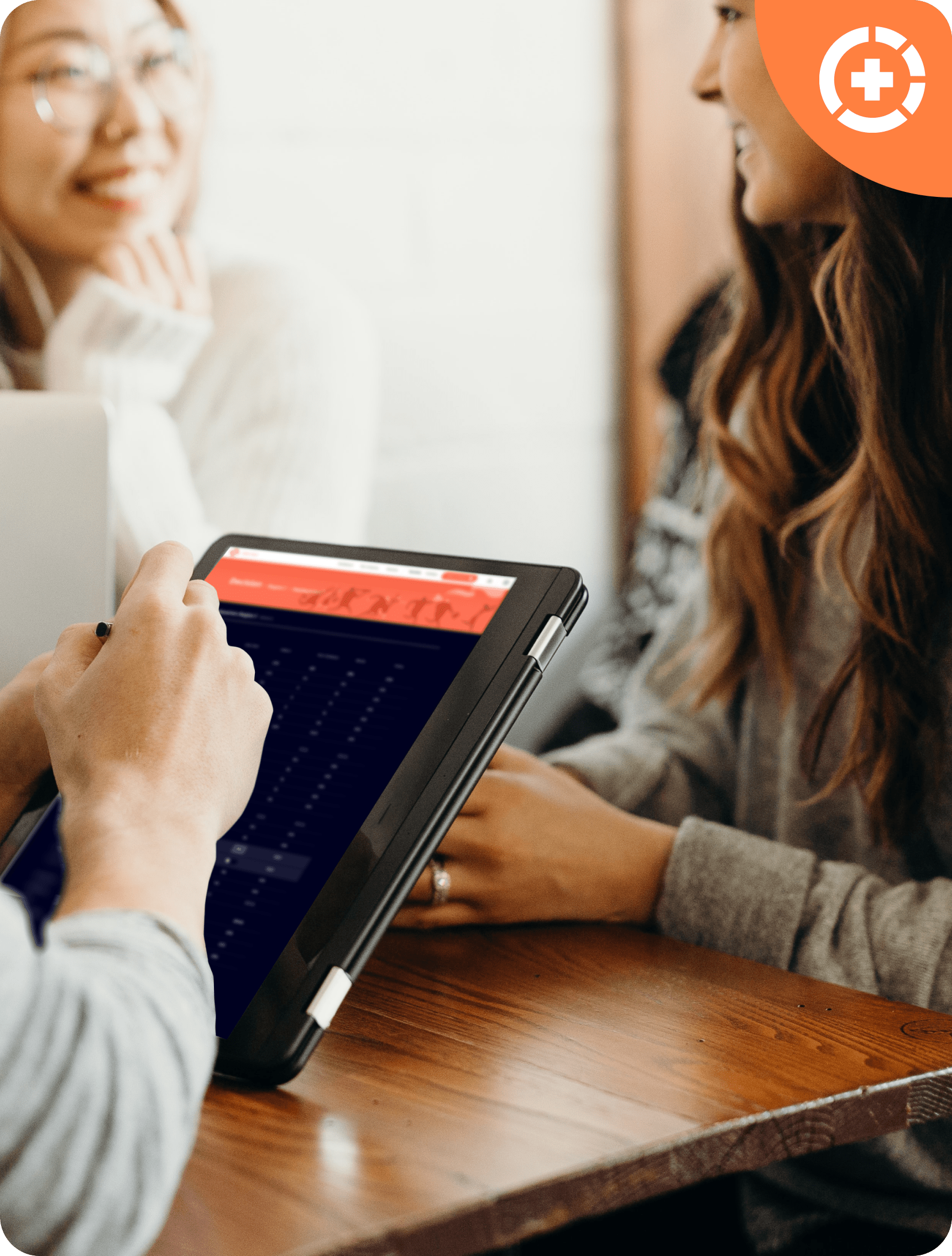 Person using a tablet with stylus while two others smile and converse across a wooden table representing the Healthcare Strategy Simulation.