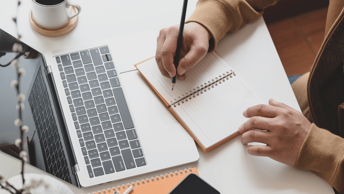 Person writing with a pencil in a spiral notebook beside an open laptop on a white desk with a coffee cup and smartphone.