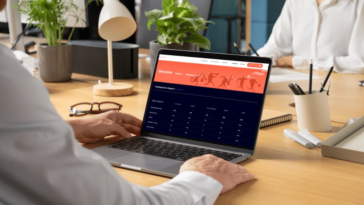 Person in white shirt working on a laptop displaying a sales report of the Healthcare Strategy Simulation on a wooden desk with office supplies and plants.