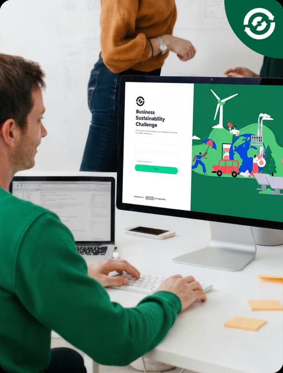 Man in green sweater typing on a keyboard at a desk with a monitor displaying a Business Sustainability Challenge login page and illustrations of wind turbines and eco-friendly activities.