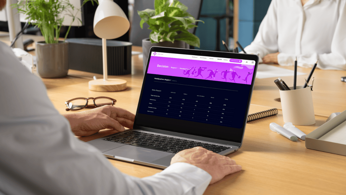 Person in white shirt working on a laptop displaying a sales report dashboard with purple header on a wooden desk with glasses, plant, lamp, and office supplies.