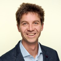 Smiling man with short brown hair wearing a blue blazer and light blue shirt against a light background.