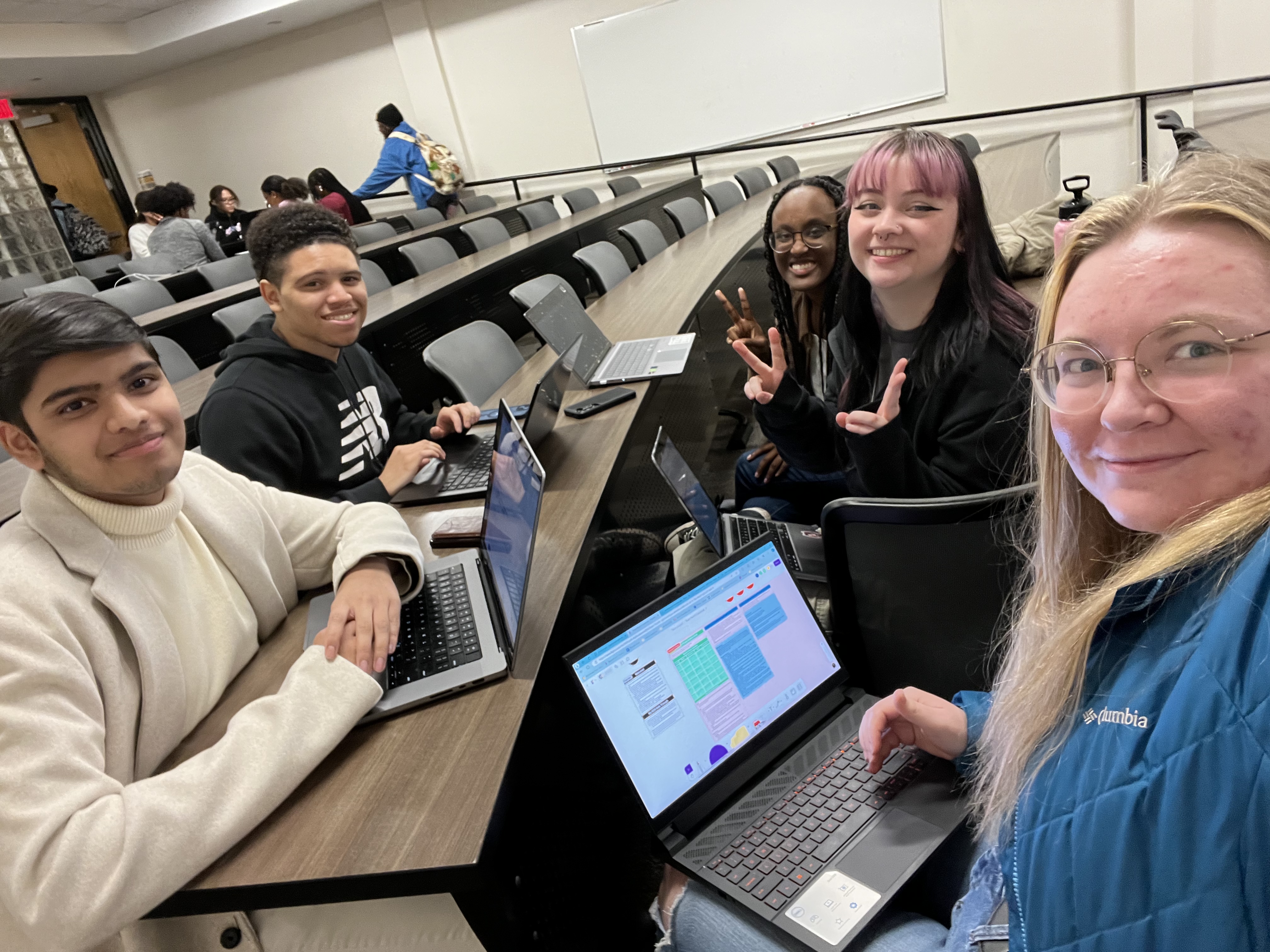 Five college students sitting with laptops around a table in a classroom, smiling at the camera, with one student making peace signs.