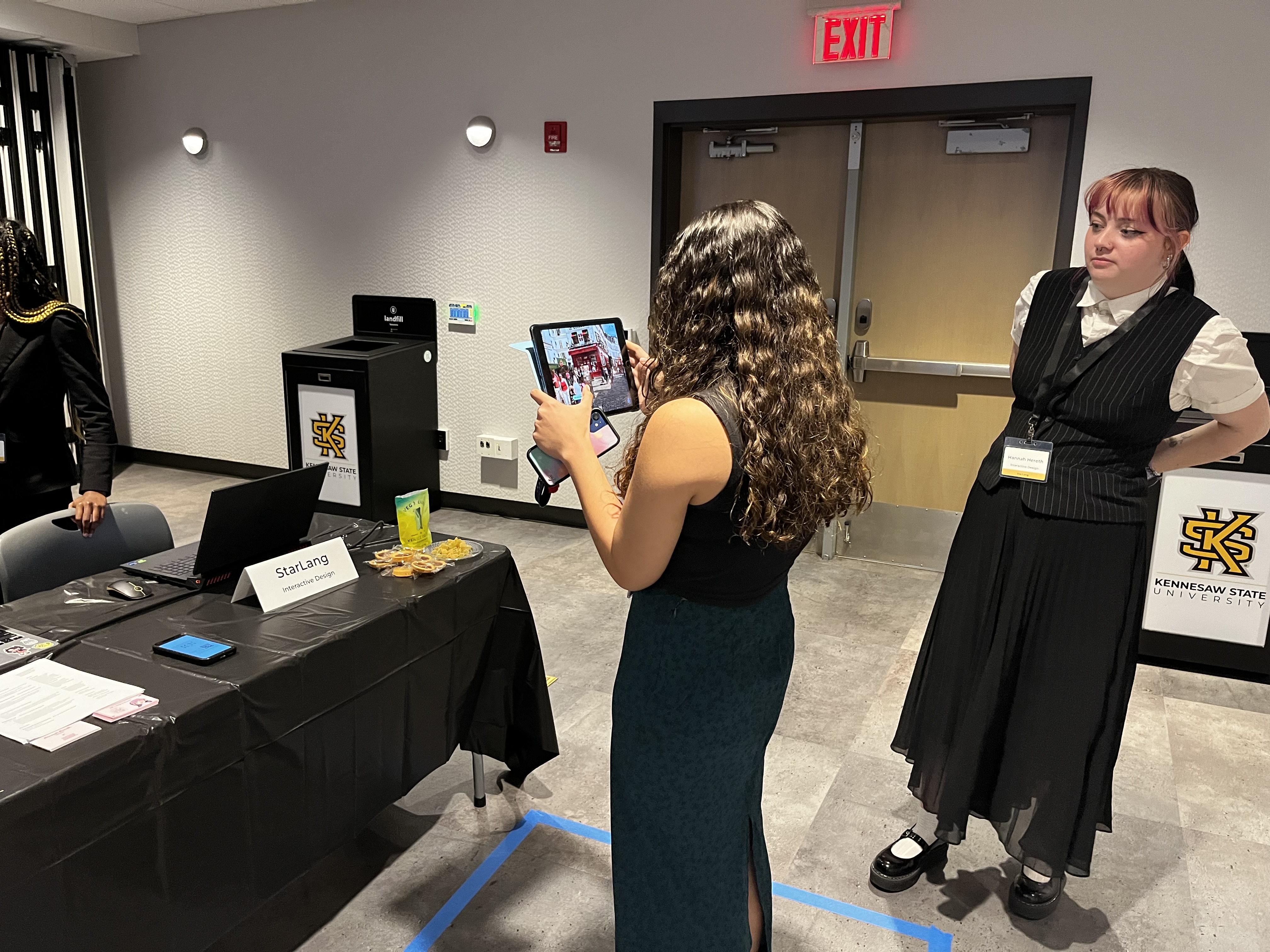 Two women at a Kennesaw State University event; one woman with long curly hair is holding a tablet showing a red phone booth, while another woman watches.