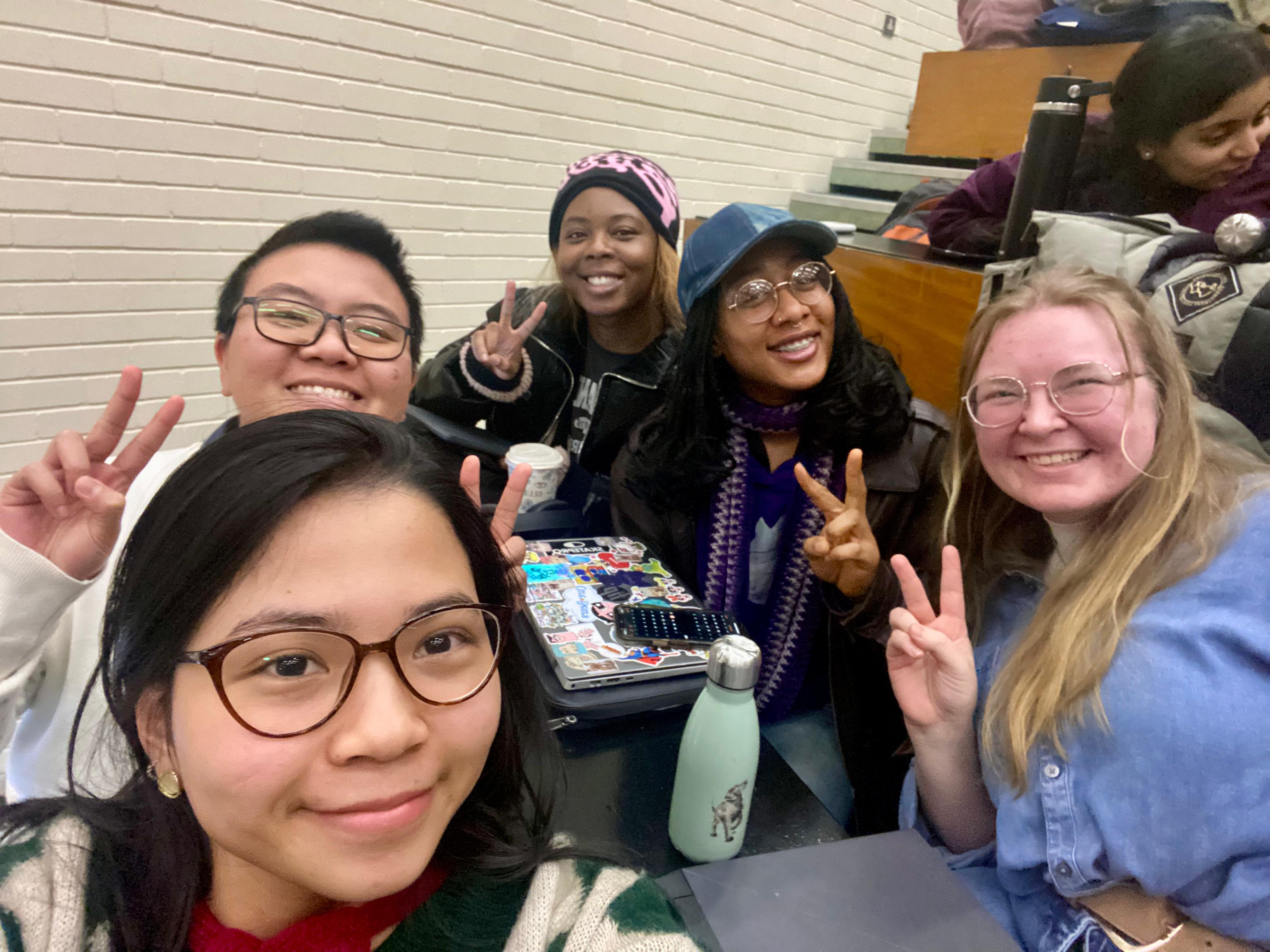 Group of five smiling young people sitting closely together, making peace signs, with laptops and water bottles on the table in a classroom setting.