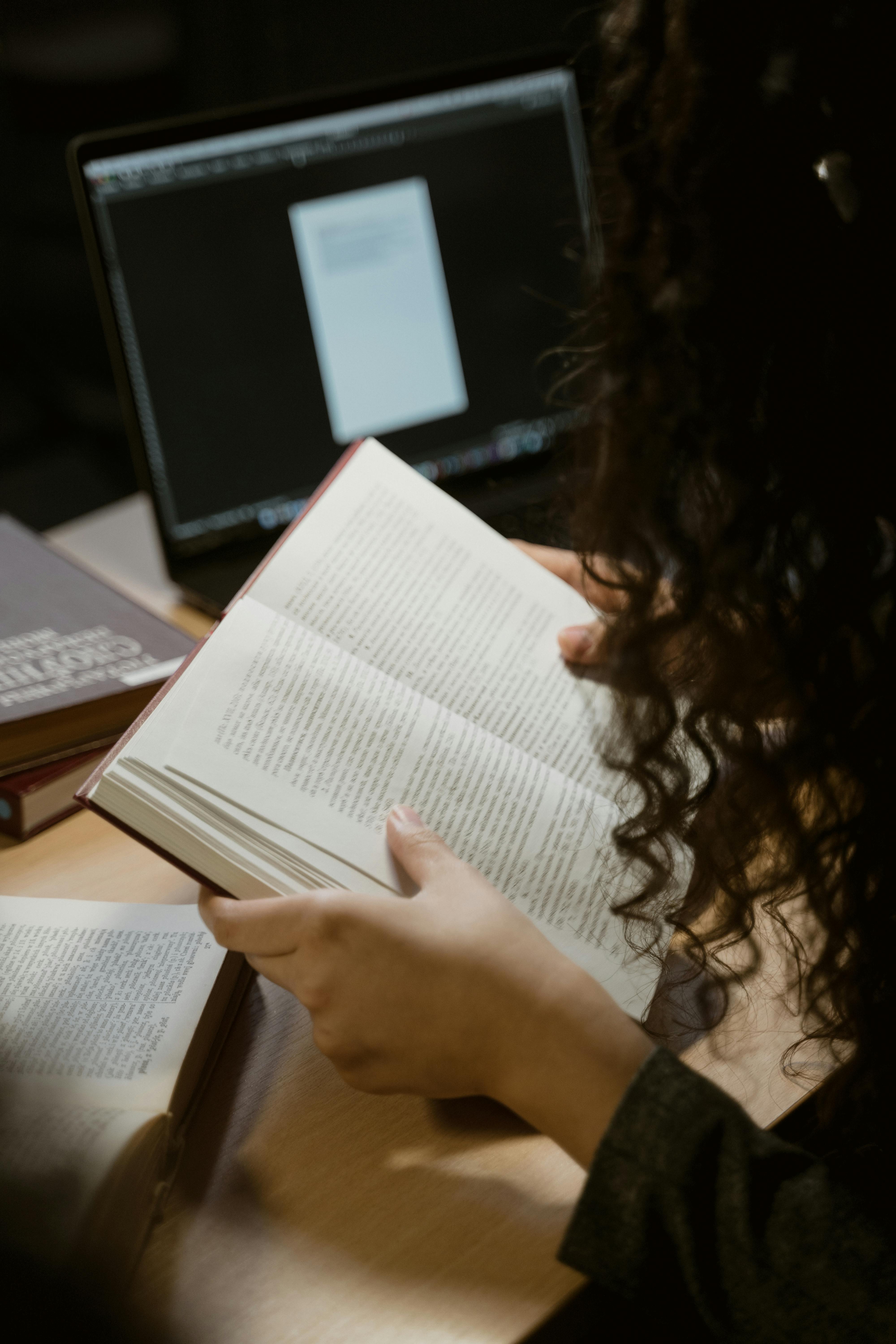 Person with curly hair reading an open book at a desk with books and a laptop in the background.
