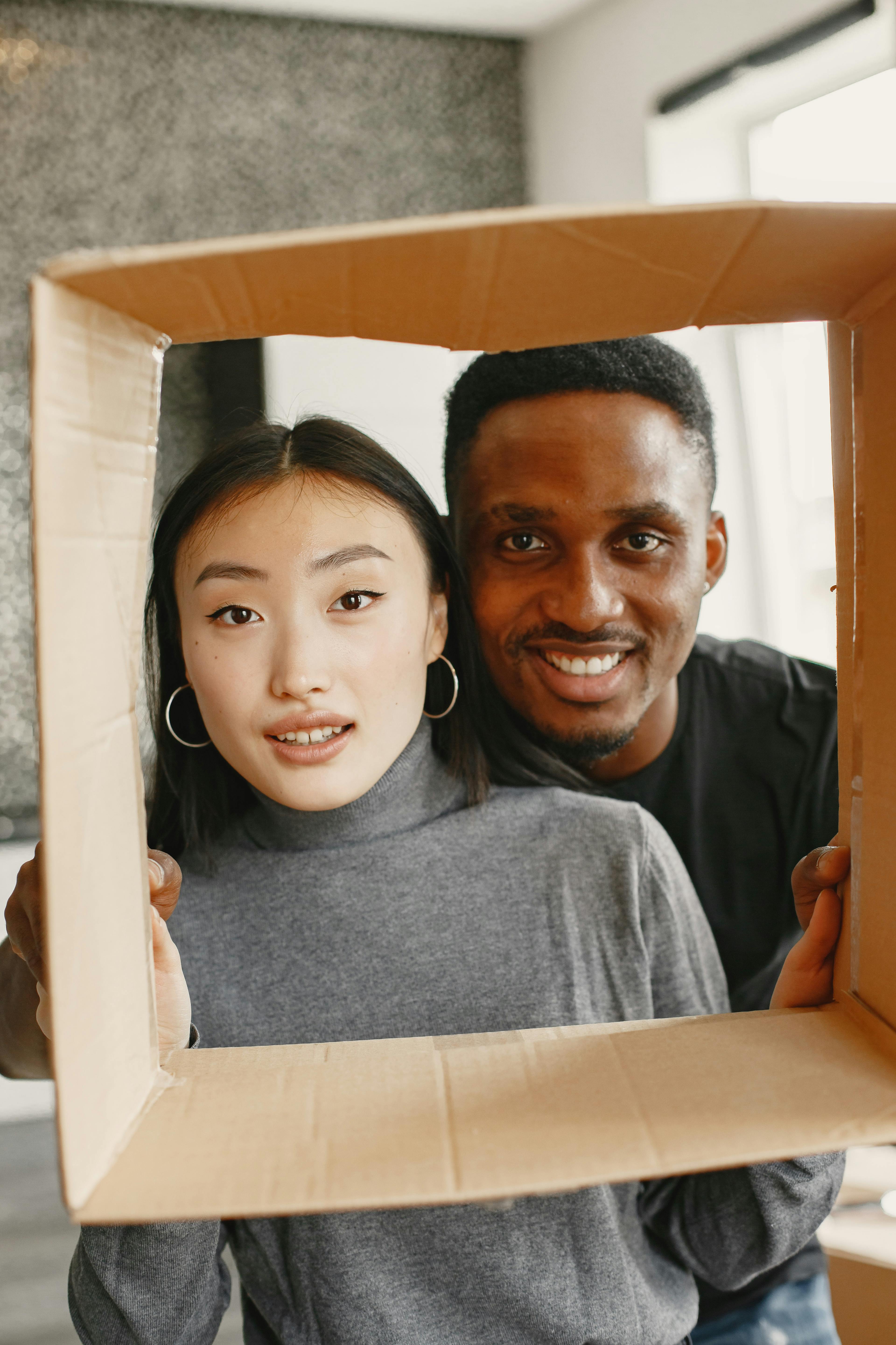 A smiling couple framed inside a large cardboard box, with the woman wearing a gray sweater and hoop earrings and the man behind her wearing a black shirt.