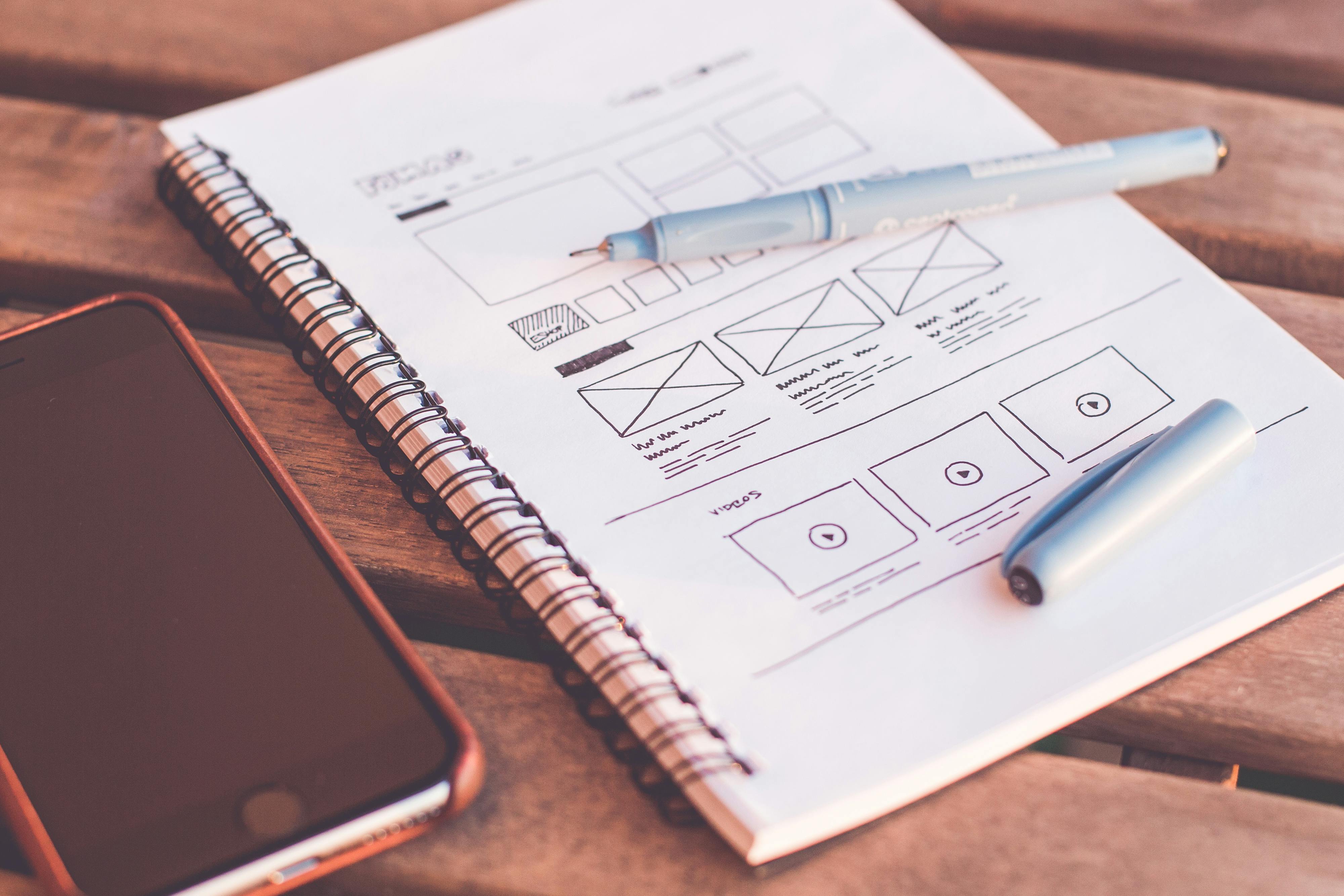 Notebook with wire-bound pages showing hand-drawn website wireframe sketches, a light blue pen with its cap off lies on the paper, and a smartphone is placed nearby on a wooden surface.