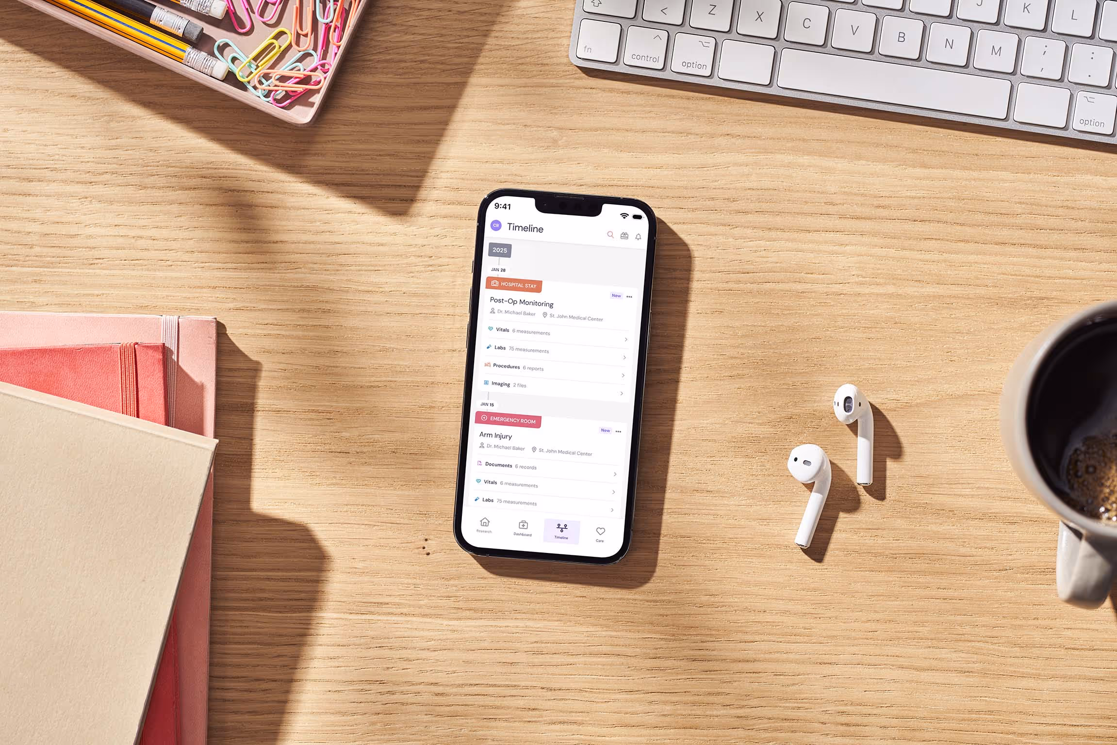 Smartphone on a wooden desk displaying a medical timeline app, surrounded by a keyboard, AirPods, a coffee cup, notebooks, and a tray with pencils and paperclips.