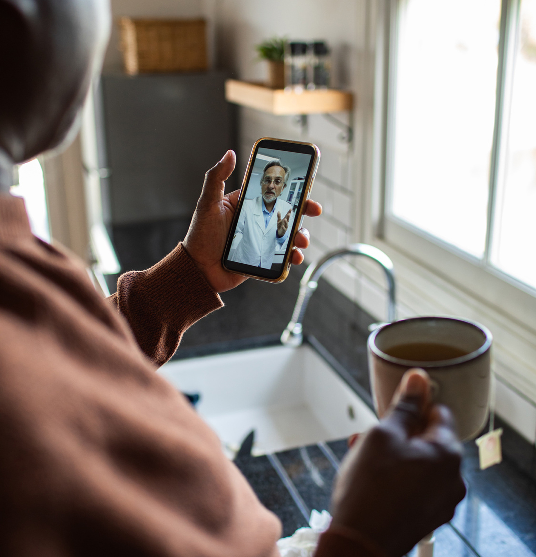 Person holding a smartphone showing a male doctor in a white coat during a video call while holding a cup of tea in a kitchen.