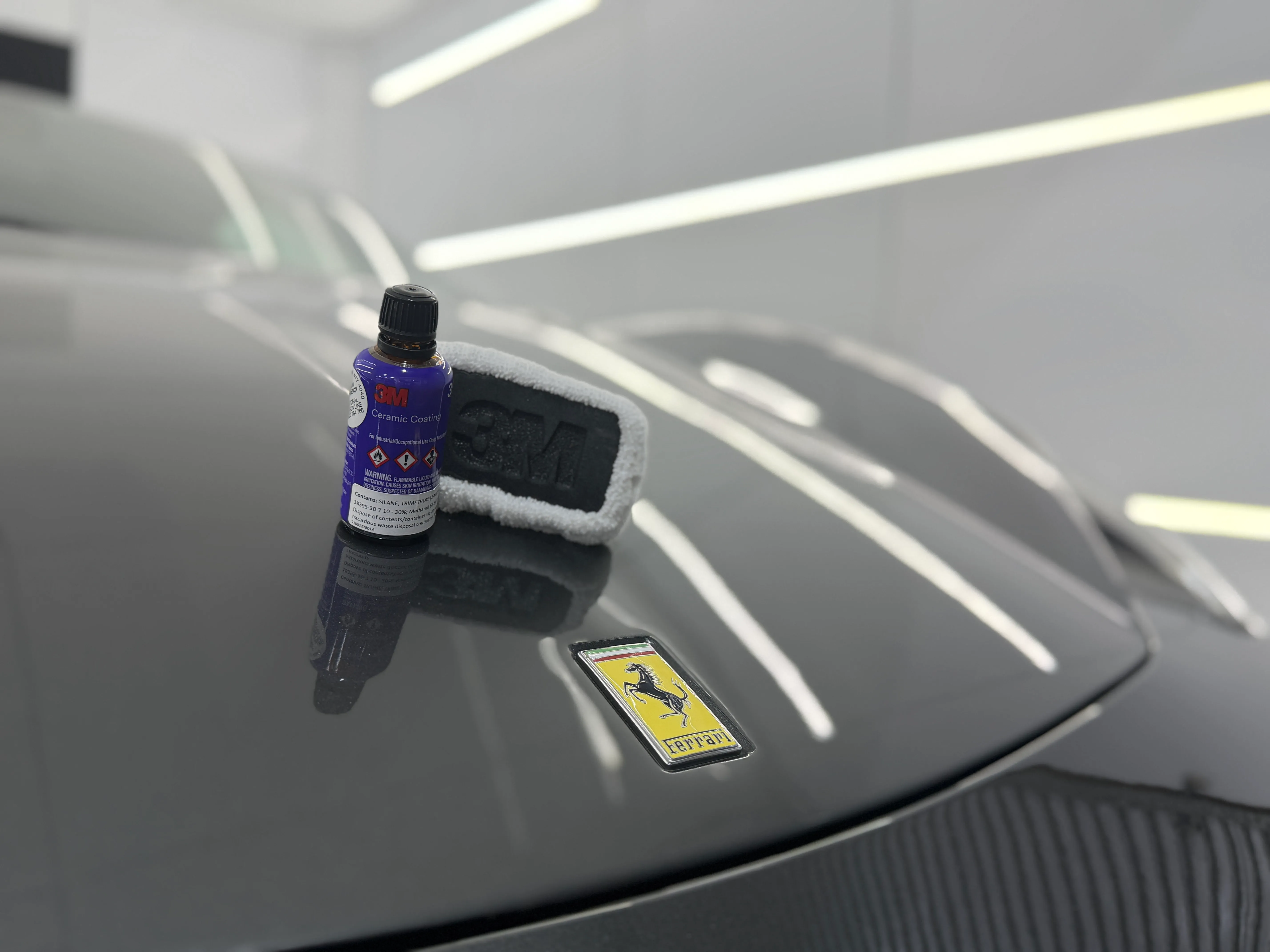 3M ceramic coating bottle and applicator pad on a glossy Ferrari hood in the workshop.