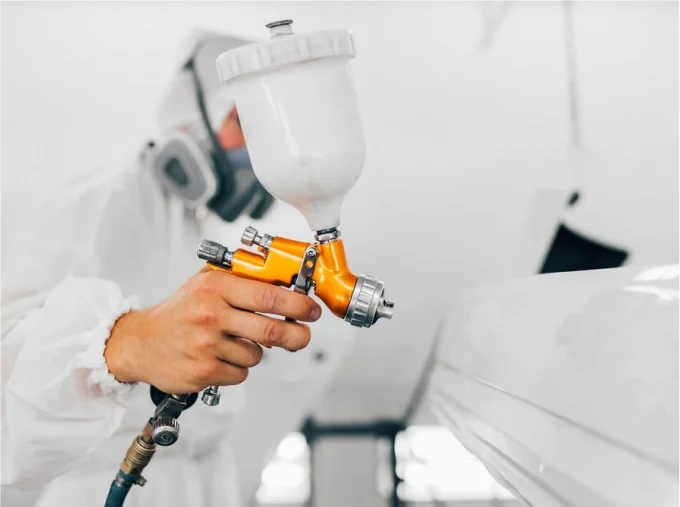 Technician in a respirator suit spraying a white vehicle panel with an orange spray gun.