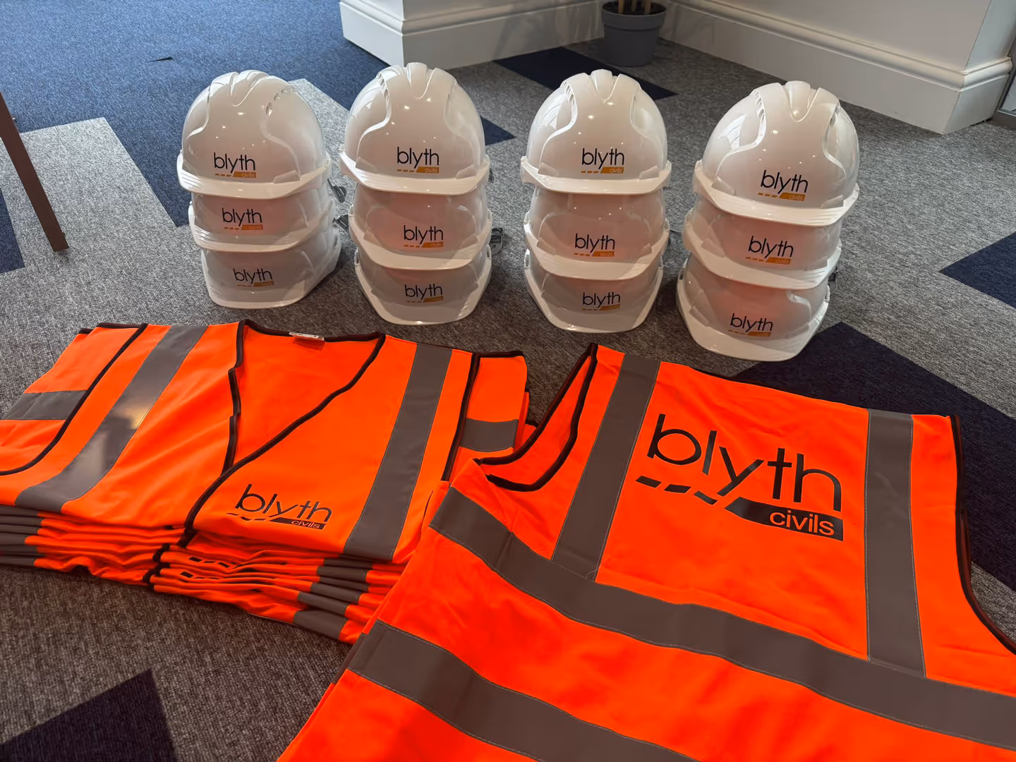 Stacks of white construction helmets and bright orange safety vests with reflective strips and 'blyth civils' logo on a carpeted floor.