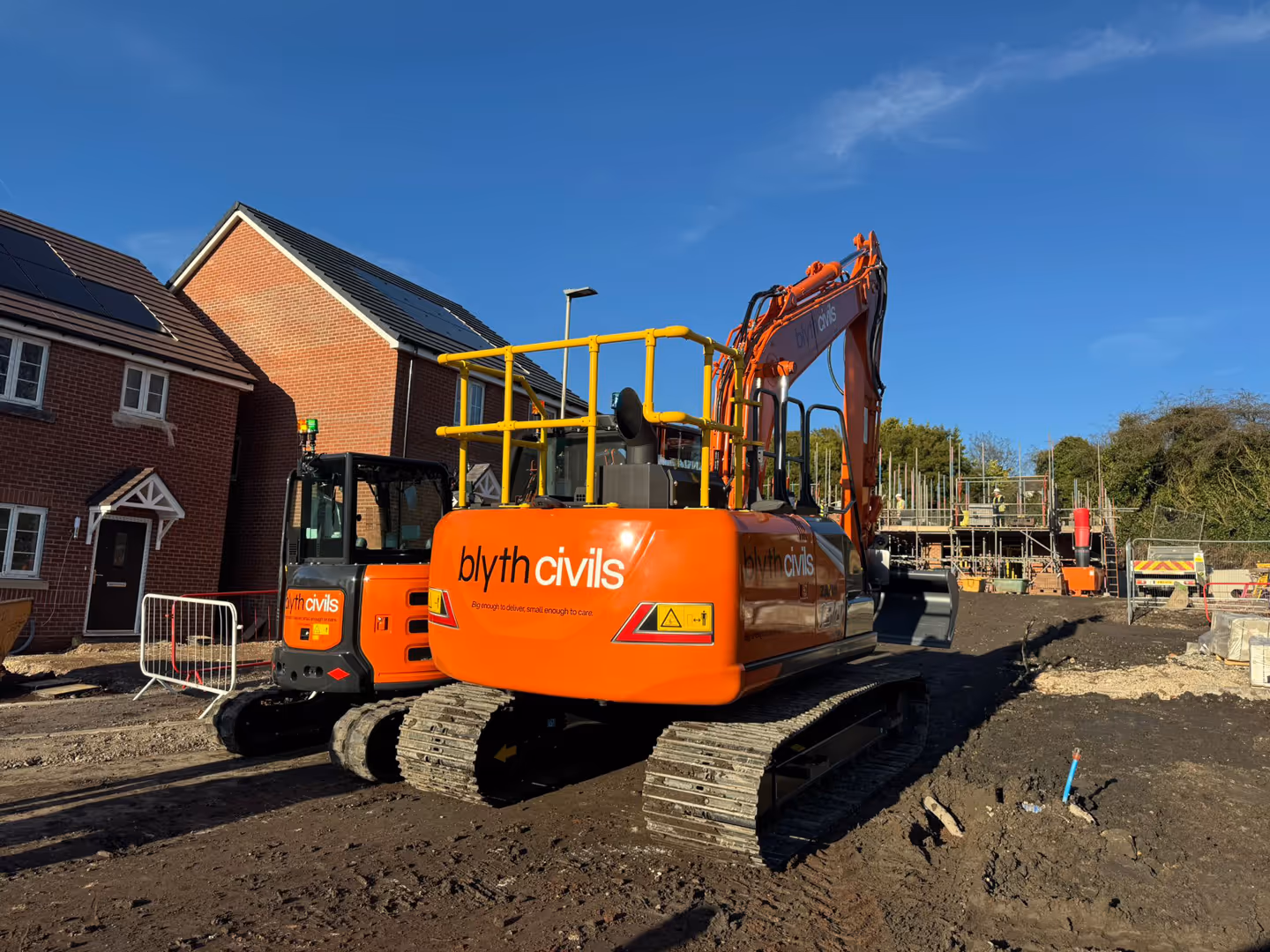 Two orange excavators with Blyth Civils branding on a construction site near brick houses under a clear blue sky.