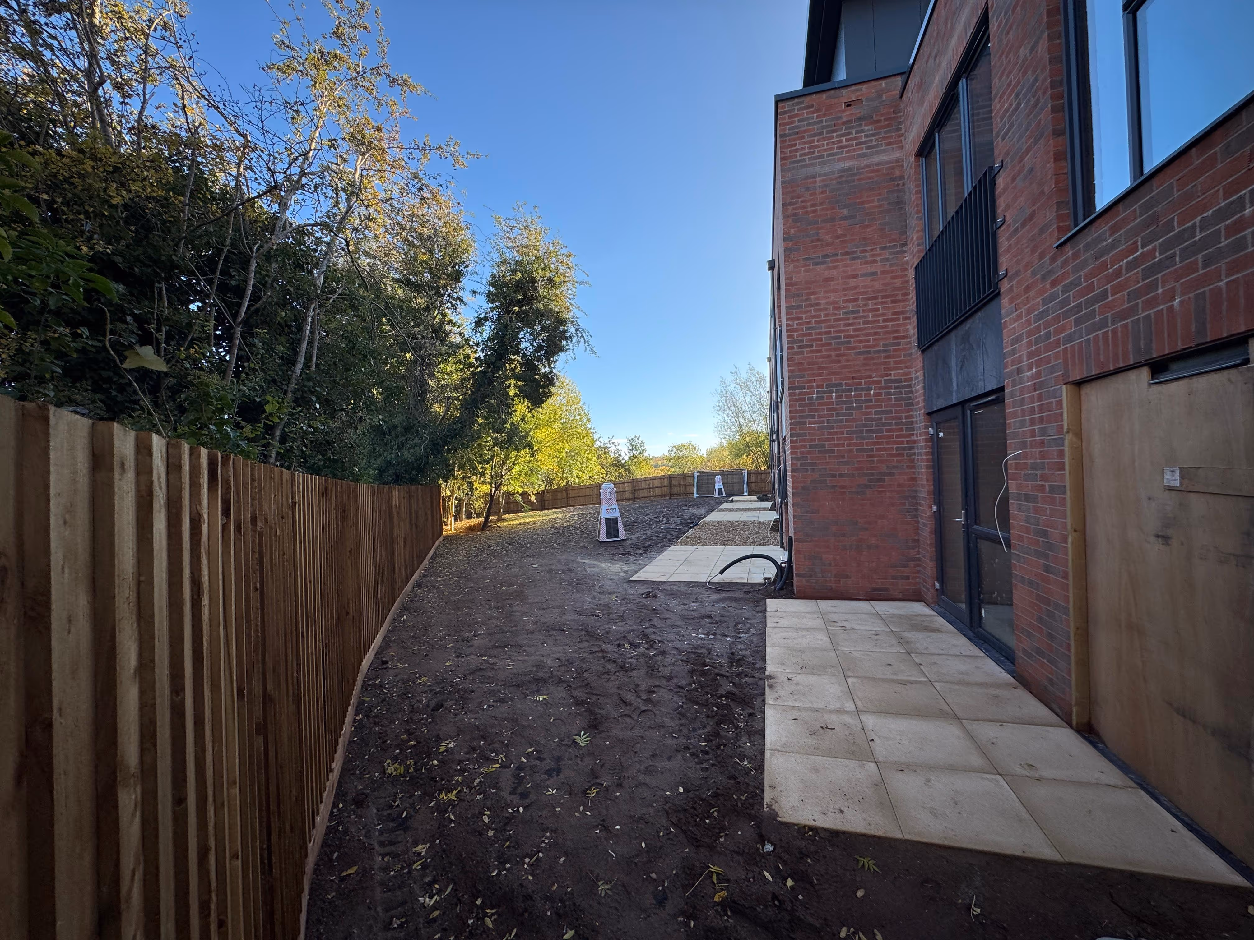 Pathway with tiled sections alongside a red brick building under clear blue sky, bordered by a wooden fence and trees.