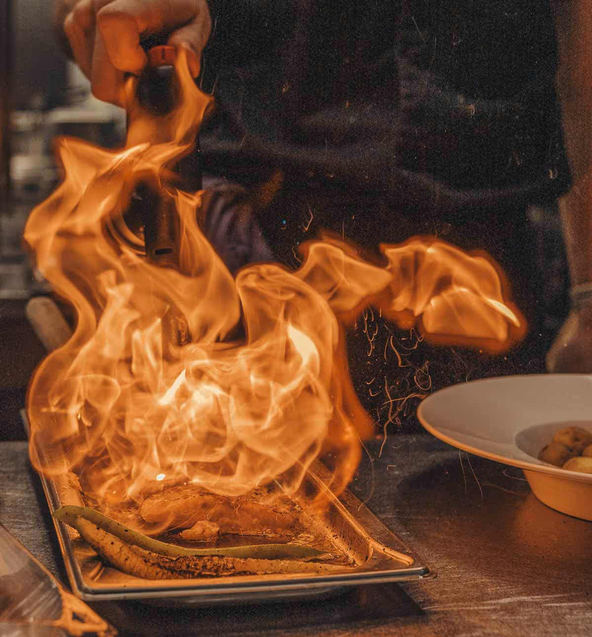 Kocken använder en gasbrännare för att flambera mat på en plåt med stark låga.