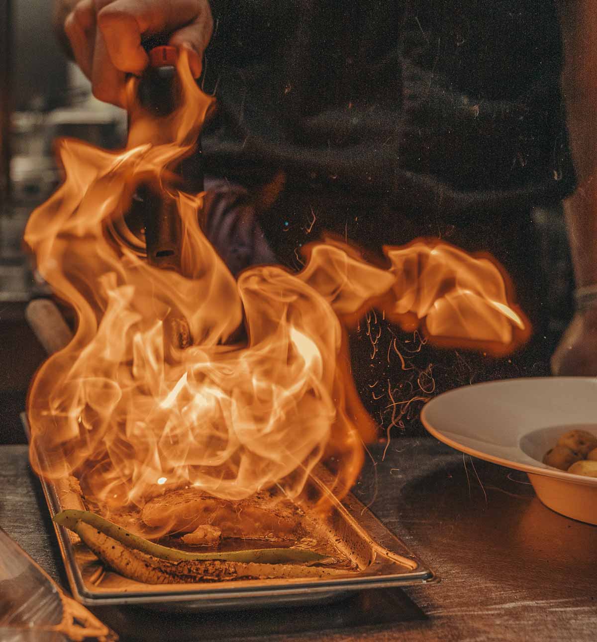 Kocken använder en gasbrännare för att flambera mat på en plåt med stark låga.