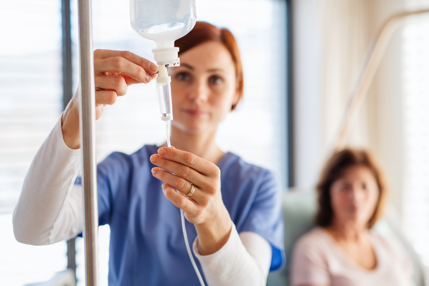Nurse adjusting IV drip with patient in background.
