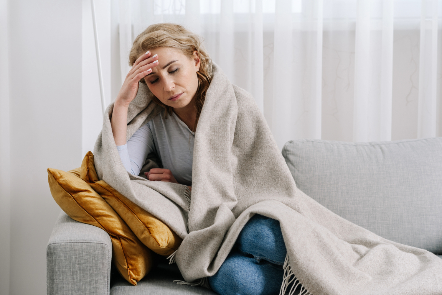 Woman in discomfort sitting on sofa, covered with blanket.