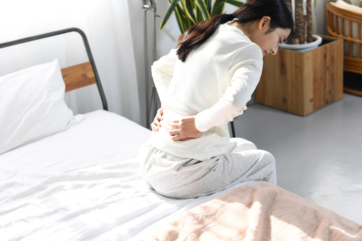 Woman sitting on bed holding lower back in pain.