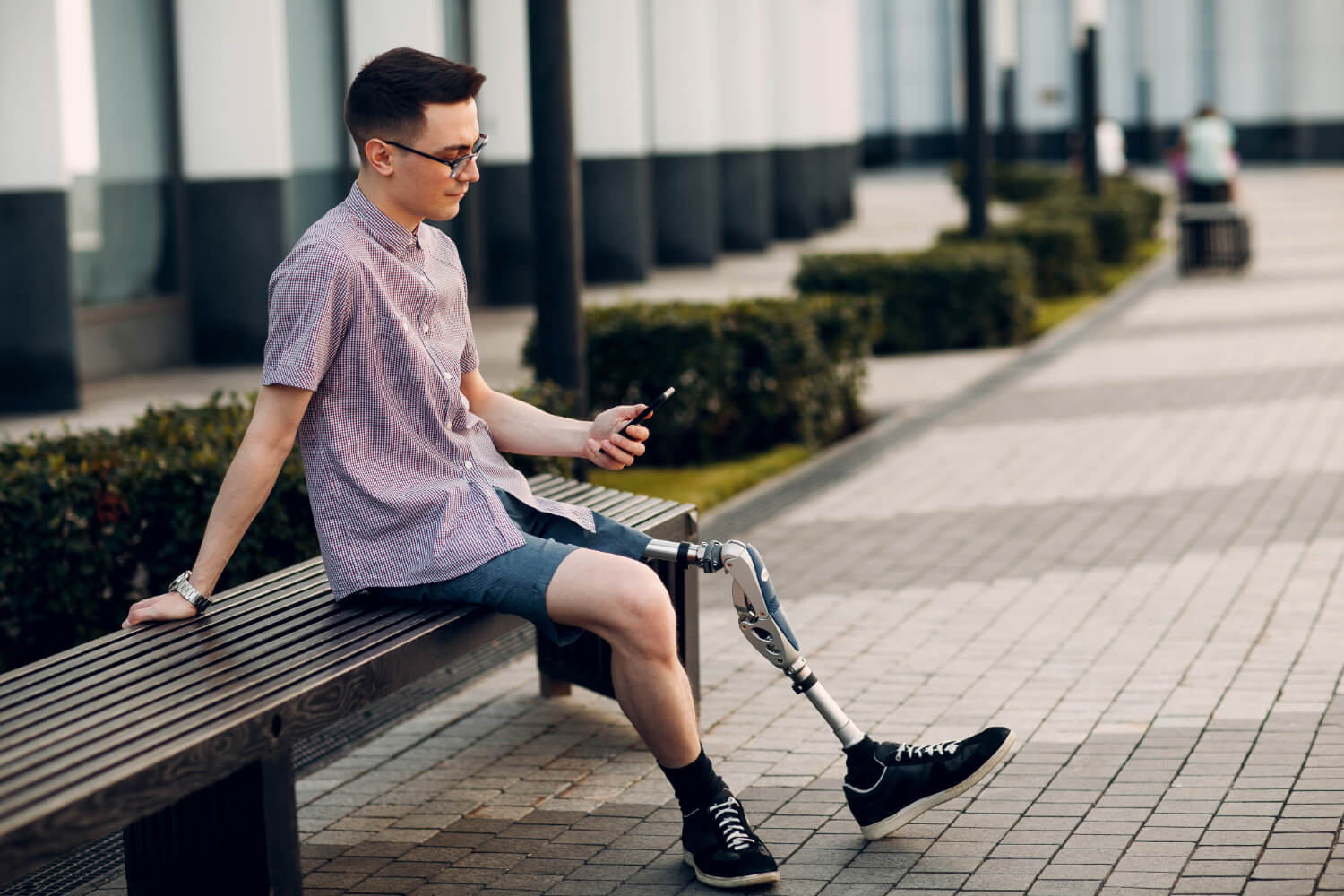 Young man with prosthetic leg using smartphone on bench outdoors