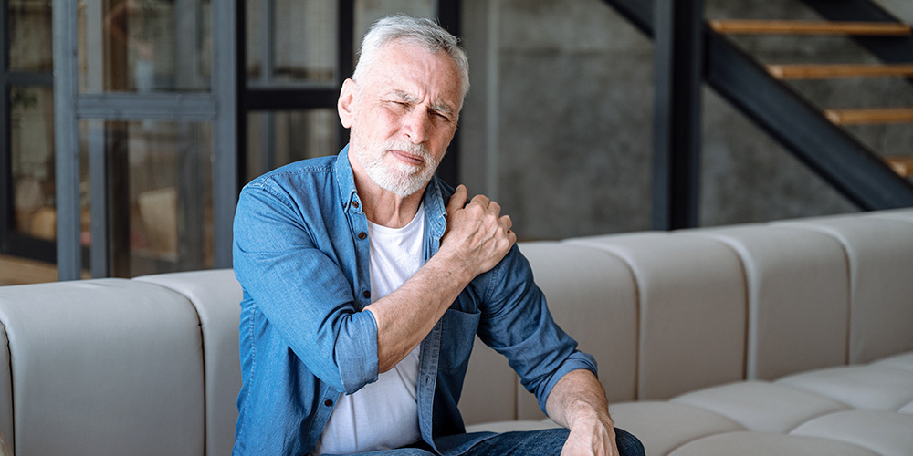 Older man in pain holding shoulder, sitting on couch.