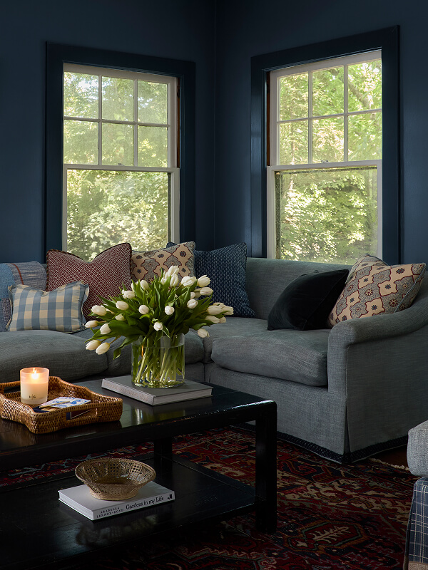 Cozy living room with gray sectional, patterned pillows, tulips in vase, candle, and large windows showing greenery.