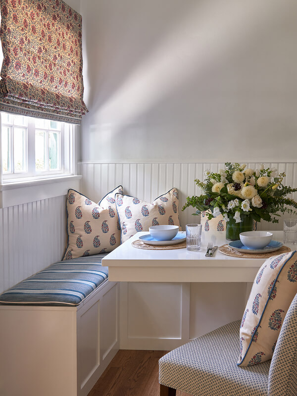 Cozy breakfast nook with floral cushions, striped bench seat, blue dishes, and a fresh flower centerpiece.