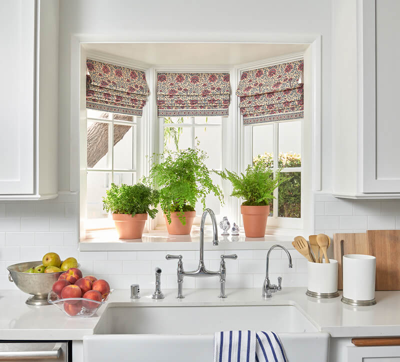 Kitchen sink with striped towel, potted plants on window sill, and fruit bowls on white counter.