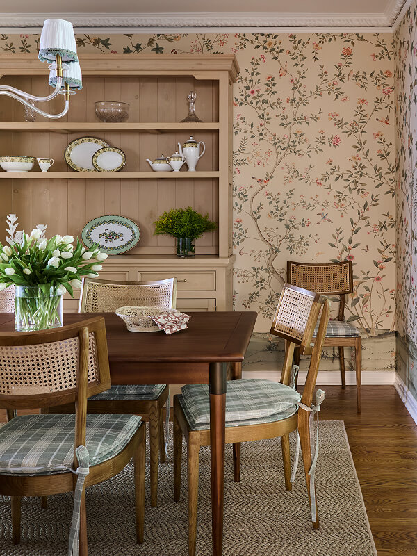 Dining room with wooden table, cane-back chairs with plaid cushions, floral wallpaper, and a shelf with china.