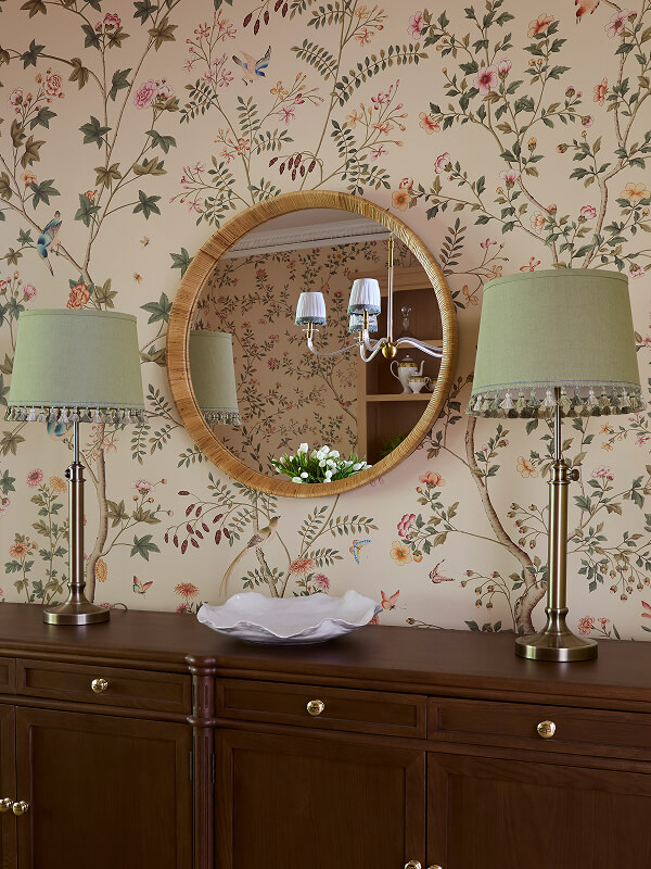 Wood cabinet with two brass lamps and a round mirror on floral wallpaper above.