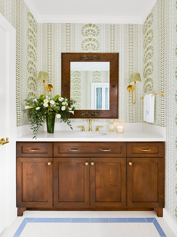 Bathroom vanity with wooden cabinets, white countertop, gold fixtures, floral wallpaper, and a flower vase.