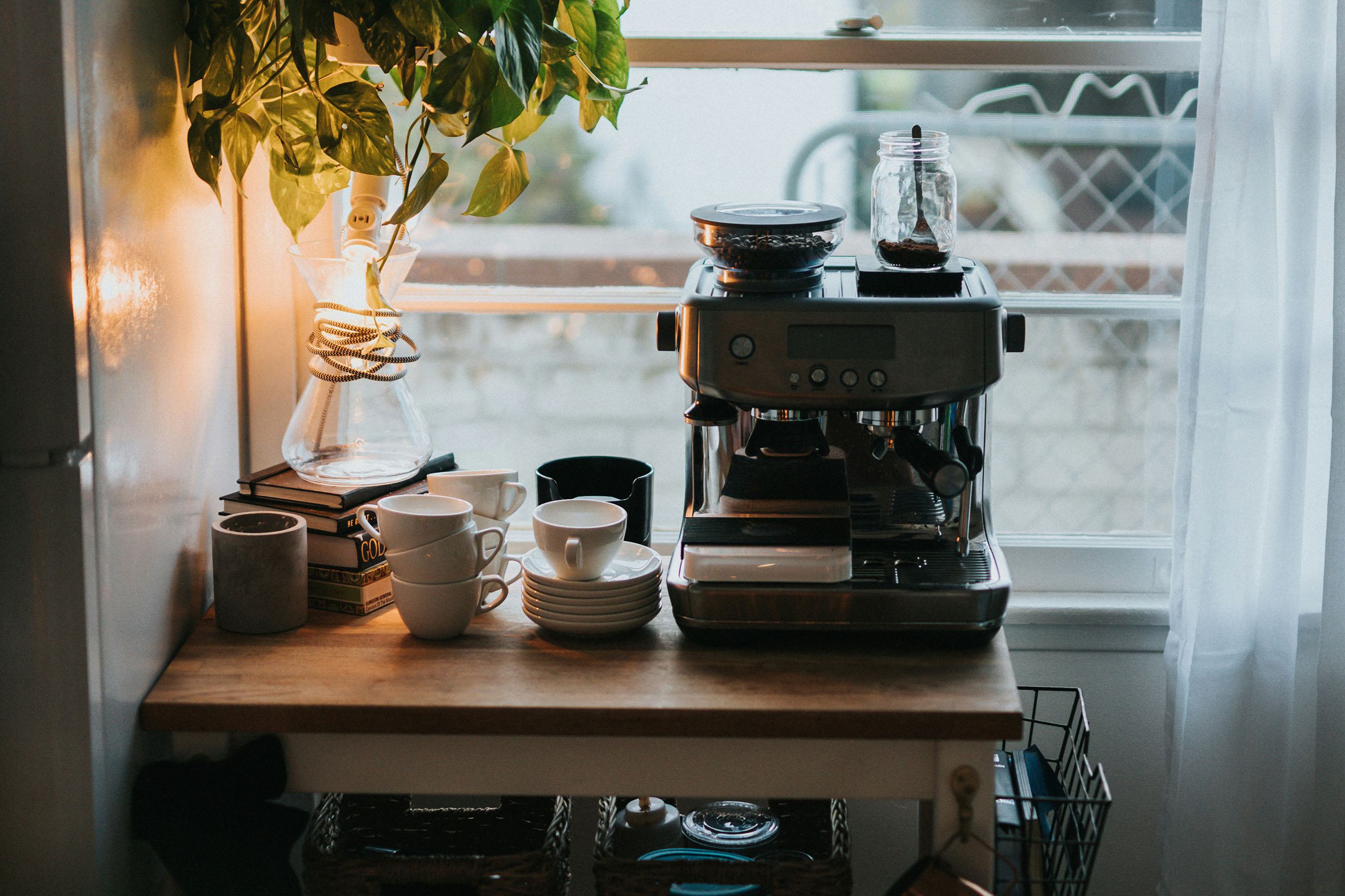 Coffee station in home kitchen next to window with small espresso machine, little mugs, plates, and a plant.