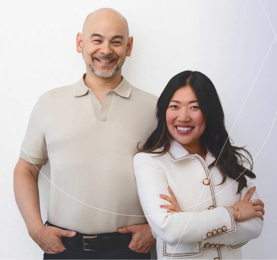 Smiling man in a beige polo shirt with hands in pockets standing next to a smiling woman with long dark hair wearing a white jacket with gold buttons and arms crossed.