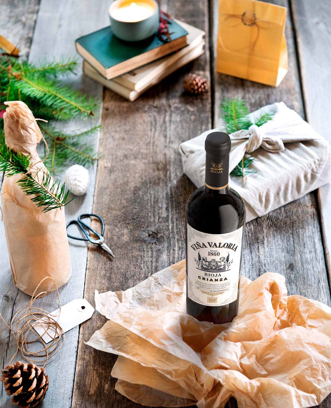 Viña Valoria Rioja Crianza bottle on rustic wood table with gift wrapping supplies, pinecones, and holiday greenery.