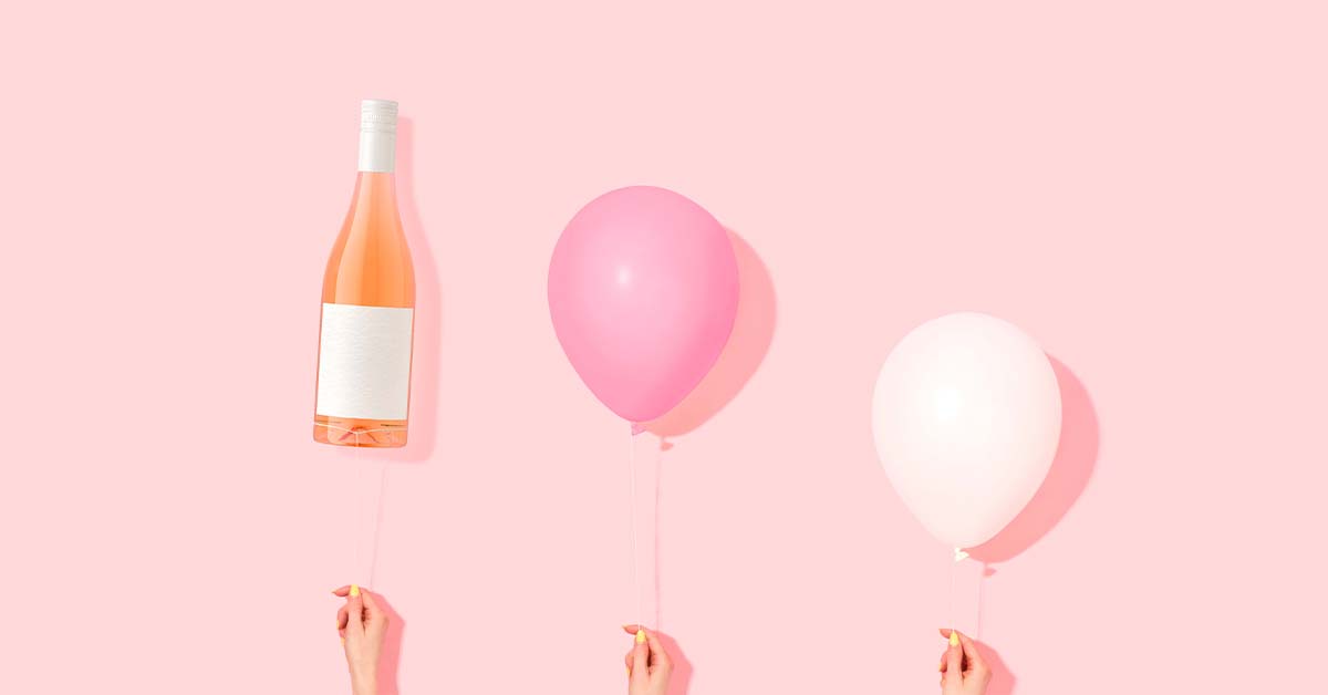 3 hands holding balloons and a bottle of rosé on a pink background