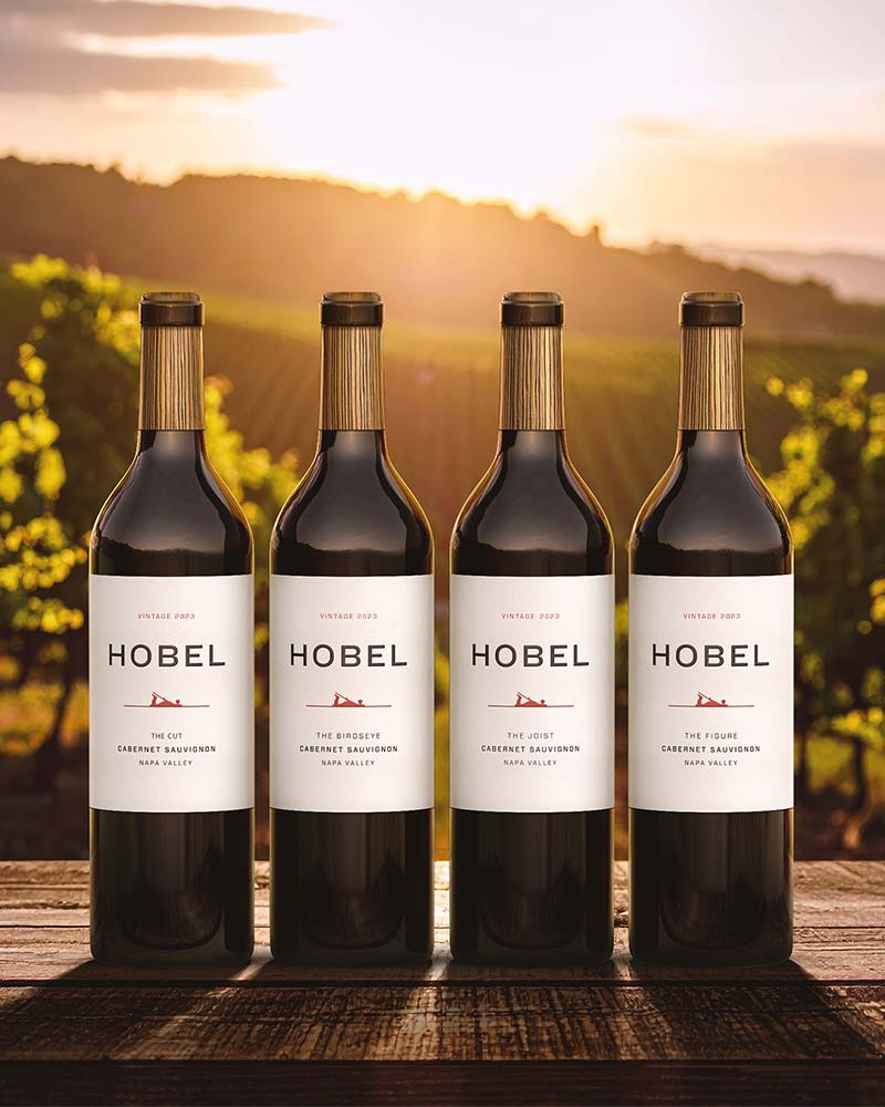 4 bottles of red wine on an outside table with a vineyard in the background at sunset