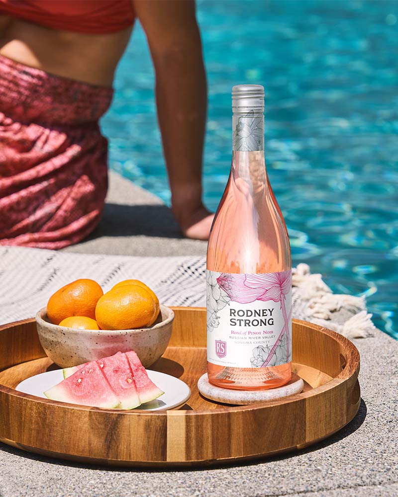 bottle of rose wine on a tray outside next to a swimming pool with woman in the background by Outshinery